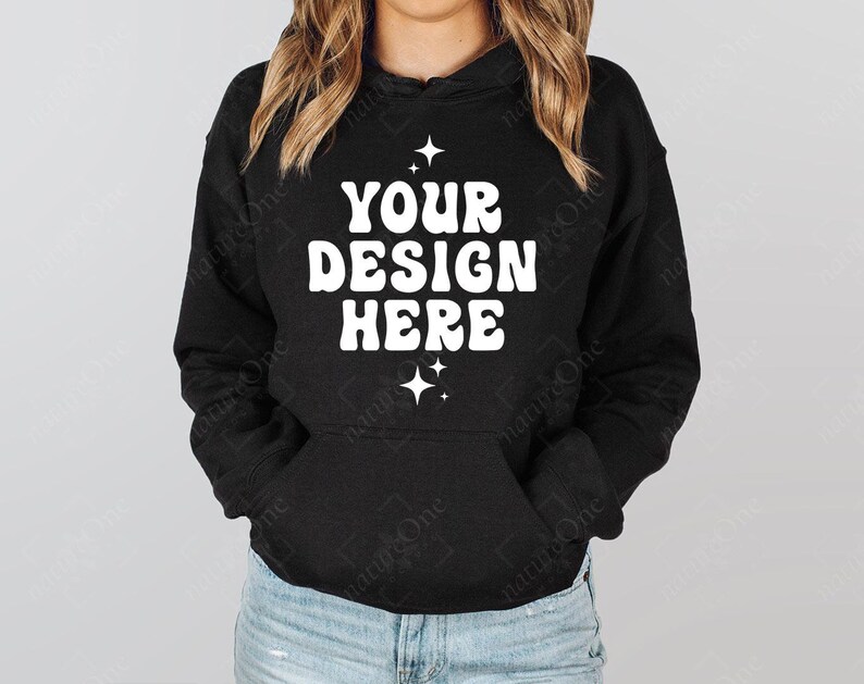 Op de afbeelding: Zwarte hoodie met de witte tekst "YOUR DESIGN HERE", met sterrenafbeeldingen. De hoodie heeft een voorzak en een capuchon met trekkoord. Het model draagt een blauwe spijkerbroek.