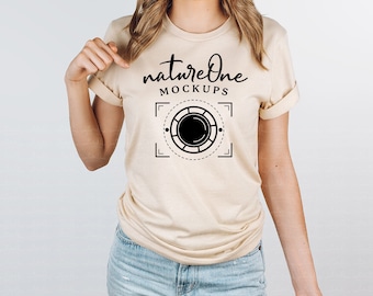 May include: A beige short-sleeved t-shirt featuring the black text natureOne MOCKUPS above a graphic of a camera lens aperture symbol enclosed in a square frame with corner brackets and dashed lines, paired with light blue denim jeans.