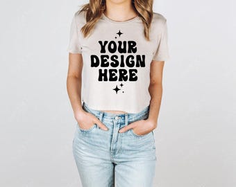 May include: A light beige short-sleeved crop top featuring the black text YOUR DESIGN HERE in a bold, wavy font, accented by small four-pointed star icons above and below the text, paired with light blue denim jeans with a button fly closure.