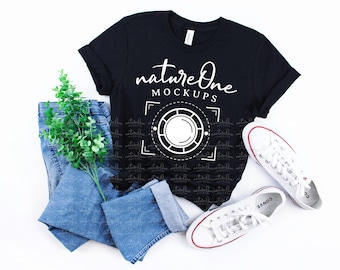 May include: A black short-sleeve t-shirt featuring the white text natureOne MOCKUPS above a circular graphic design, paired with blue denim jeans and white canvas sneakers with red trim, accented by a small green leafy plant sprig.
