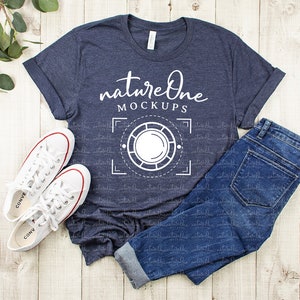 May include: A navy blue t-shirt with a white graphic design that says "natureOne MOCKUPS" and a camera icon. The shirt is laid flat on a white wooden surface with a pair of white sneakers and blue jeans.