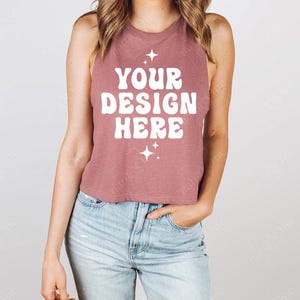 Può includere: Un top corto senza maniche color rosa polveroso con la scritta "YOUR DESIGN HERE" in bianco, insieme a grafiche a stella. Il top è abbinato a jeans di denim blu chiaro.