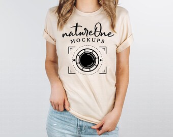 May include: A beige short-sleeved t-shirt featuring a black graphic print on the chest. The text reads natureOne MOCKUPS above a circular camera lens icon framed by dashed lines and corner brackets. The shirt has a crew neckline and rolled sleeves.