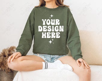 May include: An olive green crewneck sweatshirt featuring the white text YOUR DESIGN HERE in a bold, rounded font, accented by small four-pointed star graphics above and below the text, worn with light blue denim shorts and white socks.