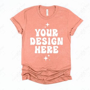 May include: A coral-colored t-shirt with rolled-up sleeves. The shirt features the text "YOUR DESIGN HERE" in a white, retro font, with starburst graphics above and below the text. The shirt is made of a soft material.