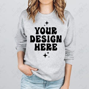 Puede incluir: Sudadera gris claro con el texto "YOUR DESIGN HERE" en negrita negra, con una fuente retro. El dise&ntilde;o est&aacute; flanqueado por dos gr&aacute;ficos de estrellas negras. La sudadera es de cuello redondo.