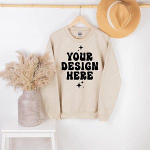 Puede incluir: Una sudadera de cuello redondo en color beige con el texto negro "YOUR DESIGN HERE" centrado en el pecho, acentuado por pequeños iconos de estrellas de cuatro puntas encima y debajo de las letras. La prenda cuelga de una percha de madera.