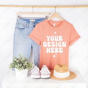 Op de afbeelding: Een perzikkleurig T-shirt met korte mouwen en de witte tekst YOUR DESIGN HERE met vierpuntige sterren, hangend aan een houten kledinghanger naast een lichtblauwe spijkerbroek, witte sneakers en een strooien hoed met een witte band.