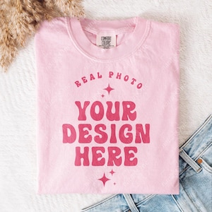 May include: A light pink crewneck t-shirt with a Comfort Colors label at the inner neckline. The front features dark pink text that reads "REAL PHOTO YOUR DESIGN HERE" in a wavy font, accented by four-pointed star graphics above and below.