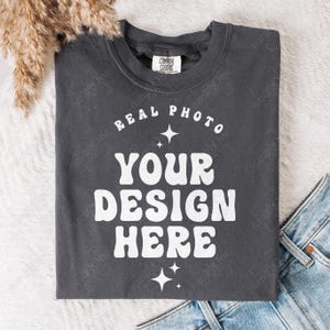 May include: A charcoal gray crew neck t-shirt featuring white screen-printed text that reads "REAL PHOTO YOUR DESIGN HERE" in a bold, wavy font. The design is accented with small four-pointed star icons above and below the main text.
