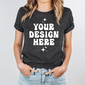 Puede incluir: Una camiseta de manga corta en color gris marengo oscuro con el texto blanco "YOUR DESIGN HERE" en una fuente redondeada y negrita, centrado en el pecho. El diseño está decorado con pequeñas estrellas de cuatro puntas arriba y abajo.