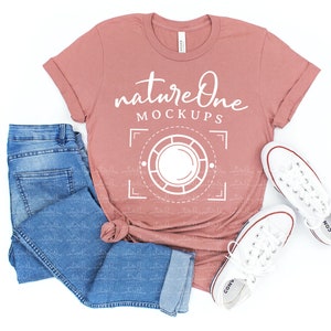 May include: A dusty rose t-shirt with the text "nature one MOCKUPS" in white. The t-shirt is laying on a white surface with a pair of blue jeans and white sneakers.