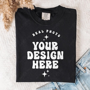 May include: A black crewneck t-shirt featuring white text that reads REAL PHOTO YOUR DESIGN HERE, arranged in a curved and stacked layout with small four-pointed star accents. The shirt has a Comfort Colors brand label inside the neckline.