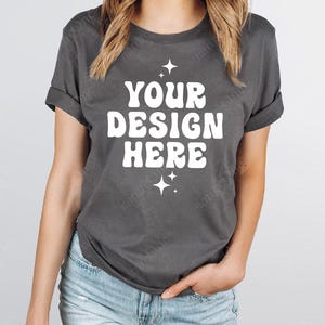 May include: A dark gray t-shirt with rolled-up sleeves. The shirt features the text "YOUR DESIGN HERE" in a white, retro font, with starburst graphics above and below the text. The model is wearing blue jeans.