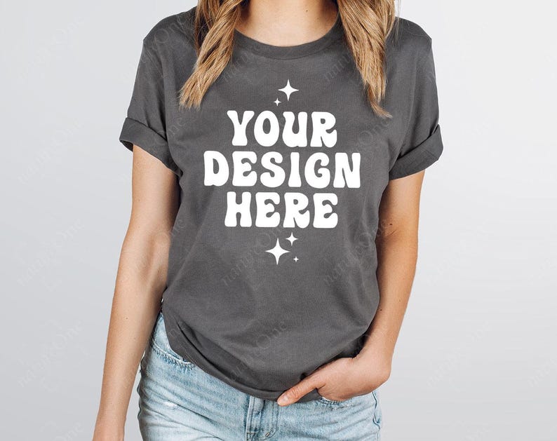 May include: A dark gray t-shirt with rolled-up sleeves. The shirt features the text "YOUR DESIGN HERE" in a white, retro font, with starburst graphics above and below the text. The model is wearing blue jeans.