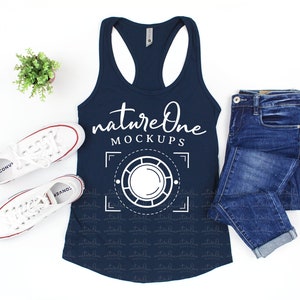 Op de afbeelding: Een marineblauw tanktop met een wit grafisch ontwerp met de tekst "nature one mockups" en een gestileerde camera lens. Het tanktop ligt plat op een wit oppervlak met een paar witte sneakers en een blauwe jeans.