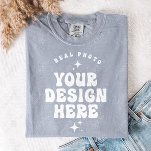 Może przedstawiać: Jasnoniebieski t-shirt z okrągłym dekoltem i białym napisem REAL PHOTO YOUR DESIGN HERE, ułożonym w zakrzywiony i warstwowy wzór z małymi czteroramiennymi gwiazdkami. Koszulka posiada metkę marki Comfort Colors przy dekolcie.