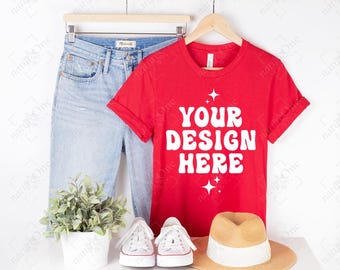 May include: A bright red short-sleeve t-shirt with the white text YOUR DESIGN HERE and star icons, displayed on a wooden hanger next to light blue denim jeans, a small potted green plant, white sneakers, and a tan straw hat with a white band.