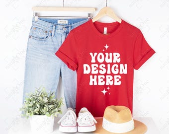 May include: A bright red short-sleeved t-shirt with the white text YOUR DESIGN HERE printed on the front, accompanied by small star icons. The shirt is displayed on a wooden hanger alongside light blue denim jeans, white sneakers, and a straw hat.