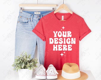 May include: A red short-sleeved t-shirt with the white text YOUR DESIGN HERE and star icons, paired with light blue denim jeans, white sneakers, a tan straw hat, and a small potted green plant, all displayed on a wooden hanger.