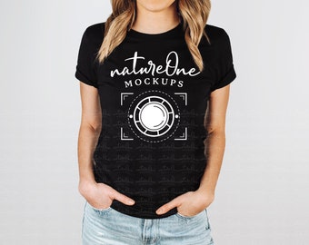 May include: A black short-sleeved t-shirt featuring white text that reads natureOne MOCKUPS above a white graphic of a camera viewfinder frame with a central circle. The shirt is paired with light blue denim jeans with hands in the pockets.