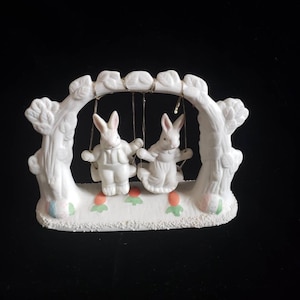 May include: White ceramic figurine of two bunnies on a swing set. The swing set is made of white ceramic and has a tree trunk design. The bunnies are wearing white and have pink noses. The swing set is decorated with green leaves and orange carrots.