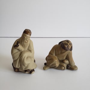 Op de afbeelding: Twee kleine, zittende beeldjes. Eén figuur is gekleed in een lichtbeige gewaad en hoed, met een voorwerp in de hand. De andere figuur heeft een vergelijkbare houding, met een bruin gewaad en baard. Beide staan op een wit oppervlak.