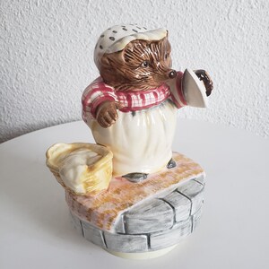May include: A ceramic musical figurine of a hedgehog wearing a red and white checkered dress and a white polka dot hat. The hedgehog is holding a small basket and a pitcher. The figurine is standing on a stone base.
