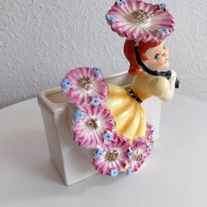 May include: A vintage ceramic planter featuring a girl with red hair and a yellow dress, adorned with pink and blue flower accents. The planter is white and has a rectangular shape. The girl is holding a black bow.