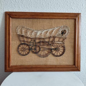 May include: A string art wall hanging featuring a covered wagon design created with dark brown and cream-colored thread on a tan burlap background, enclosed within a rectangular wooden frame with a natural medium-brown finish.
