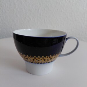 Op de afbeelding: Een marineblauwe theekop met een witte binnenkant en basis. De kop heeft een gouden decoratief patroon rond het midden. Het handvat is lichtblauw.