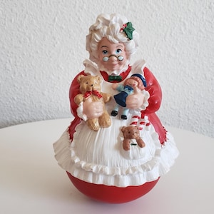May include: A ceramic Mrs. Claus figurine in a red dress with a white apron, holding a teddy bear and a doll. The figurine has a round red base and is decorated with green holly accents, perfect for holiday decor.