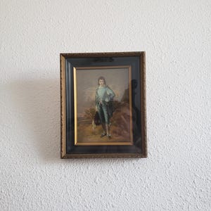May include: A framed painting of a young boy in a blue suit standing in a field. The painting is in a gold frame with a black mat.