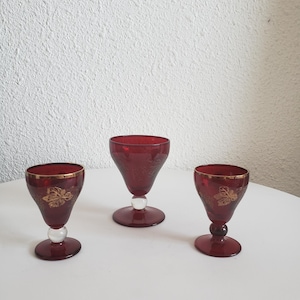 May include: Three red glass shot glasses with gold leaf accents. The glasses have a round base and a flared top.