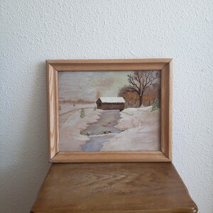 May include: A framed painting of a winter scene with a covered bridge over a stream. The trees are bare and the ground is covered in snow. The painting is in a wooden frame.