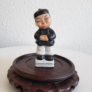 Może przedstawiać: Mała ceramiczna figurka młodego człowieka w tradycyjnym chińskim stroju, z czarną kurtką, białymi spodniami i czarnym kapeluszem. Figurka stoi na ciemnej drewnianej podstawie z dekoracyjną podstawą.