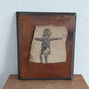 May include: Vintage framed print of a Native American archer. The print is affixed to a dark wooden board with a black border. The archer is depicted in a traditional outfit, aiming a bow and arrow.