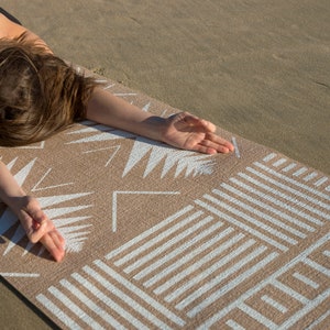 May include: A person is lying on a beige yoga mat with a white geometric pattern. The mat has a textured surface and is designed for use on the beach or outdoors.