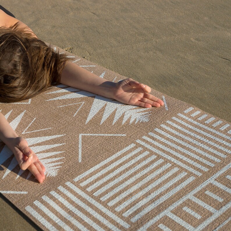 May include: A person is lying on a beige yoga mat with a white geometric pattern. The mat has a textured surface and is designed for use on the beach or outdoors.