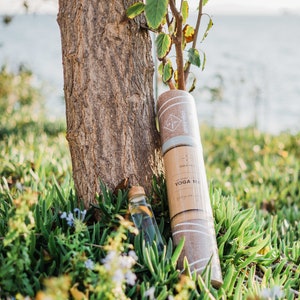 May include: A brown yoga mat with white stripes and the text "ORGANIC YOGA MAT" is rolled up and leaning against a tree trunk. A clear glass bottle with a cork top is in the foreground.