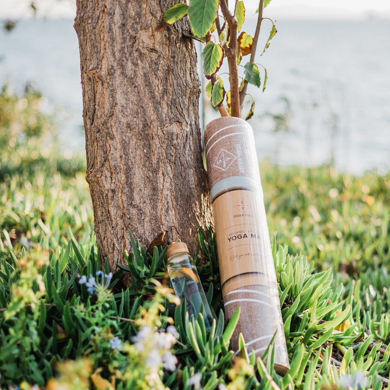 May include: A brown yoga mat with white stripes and the text "ORGANIC YOGA MAT" is rolled up and leaning against a tree trunk. A clear glass bottle with a cork top is in the foreground.