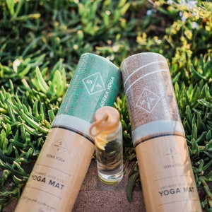 May include: Two rolled up yoga mats with brown paper packaging. The mats are made of organic jute and are eco-friendly. The text on the packaging reads "Organic Jute Eco Yoga Mat" and "Seeka Yoga".
