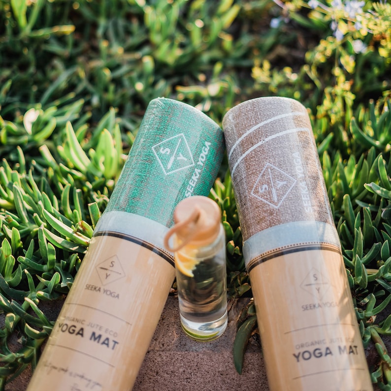 May include: Two rolled up yoga mats with brown paper packaging. The mats are made of organic jute and are eco-friendly. The text on the packaging reads "Organic Jute Eco Yoga Mat" and "Seeka Yoga".
