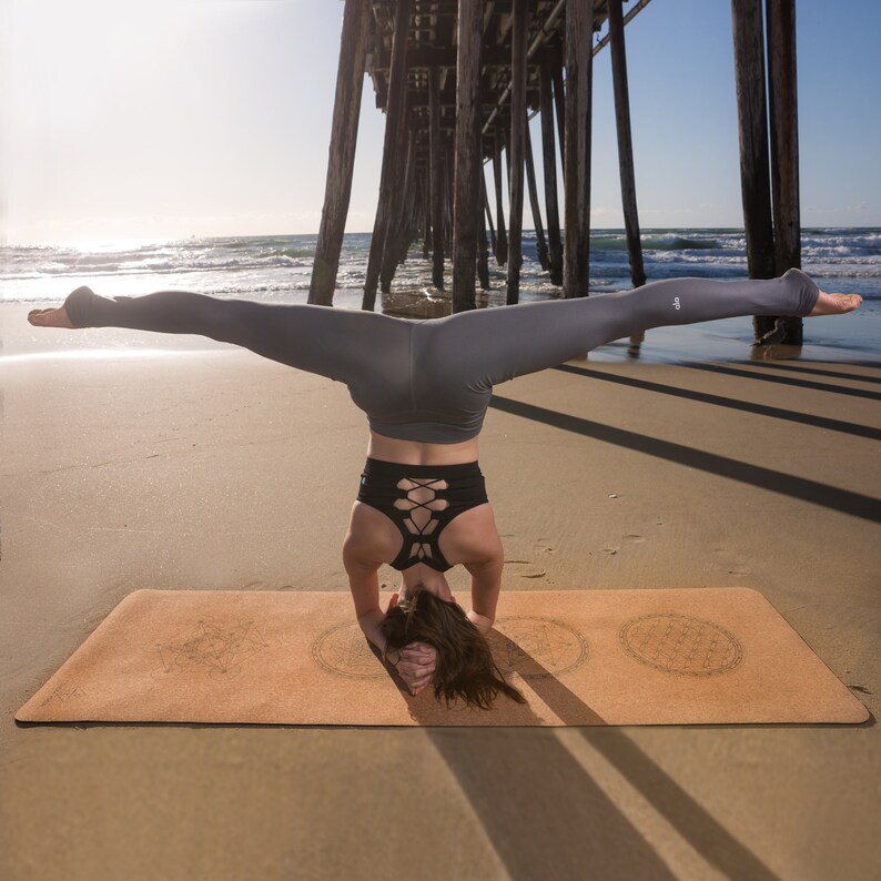 May include: A woman performs a yoga pose on a cork yoga mat with a mandala design. She is wearing a black sports bra with a criss-cross back and gray leggings. The mat is on a sandy beach with a wooden pier in the background.