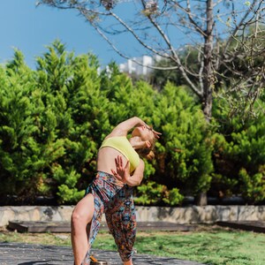 Op de afbeelding: Een vrouw in een geel tanktop en een broek met patroon voert een yogapose uit op een kurken yogamat buiten. Een zangkom staat op de mat.