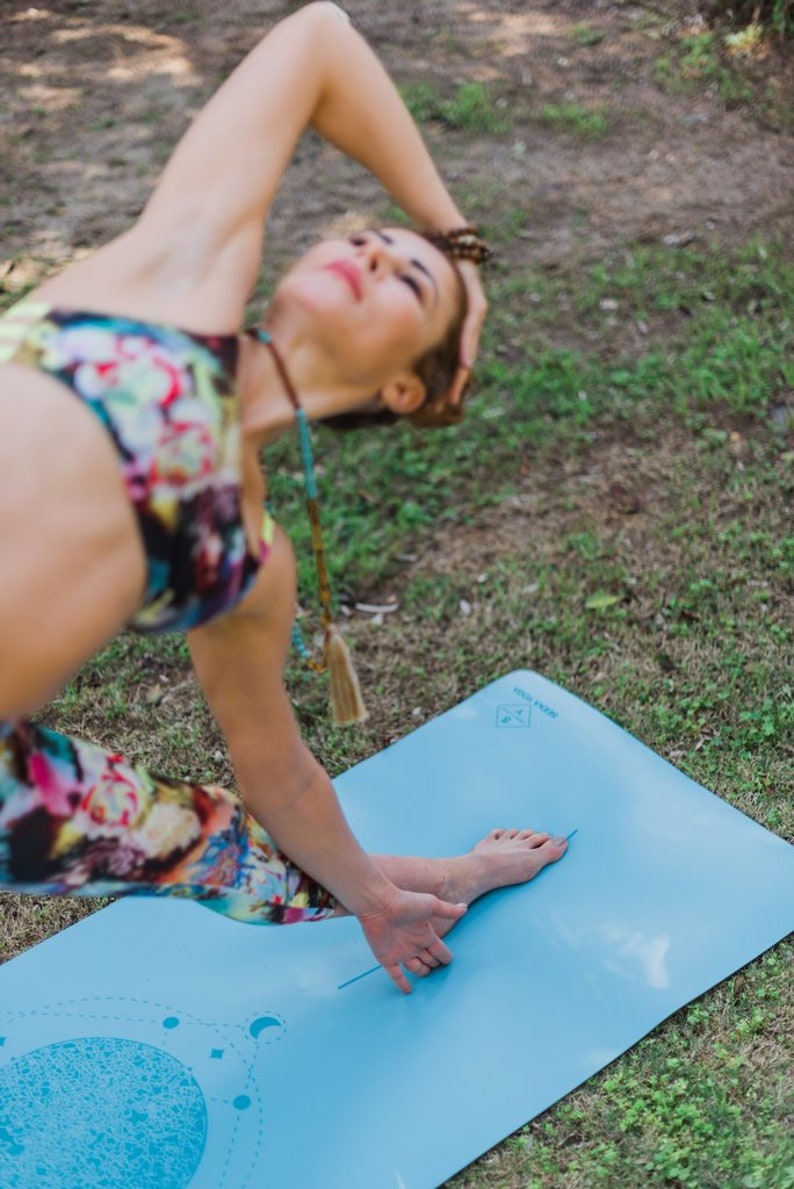 May include: A woman in colorful leggings and a sports bra practices yoga on a blue yoga mat with a moon and star design. The mat has the words "Yoga Mat" printed on it.
