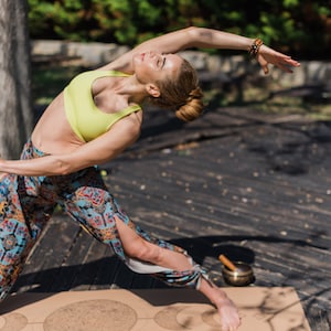 Op de afbeelding: Een vrouw in een gele sportbeha en een broek met patroon voert een yogapose uit op een kurken yogamat met een maan- en sterrendesign. Een zangkom staat op de mat.