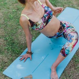 May include: A woman in a floral sports bra and leggings is doing a yoga pose on a blue yoga mat. The mat has a white line down the center and the words "Seeka Yoga" printed on it. A beaded necklace is lying on the mat.