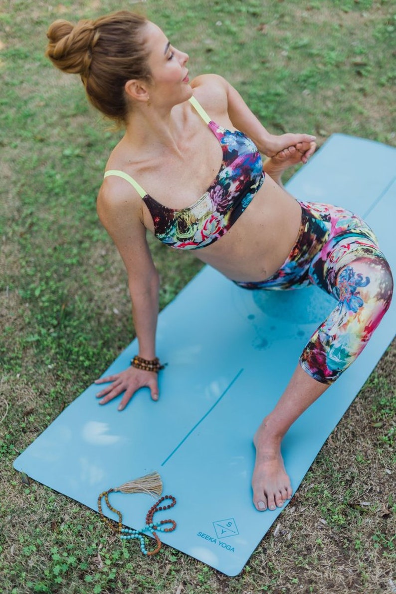 May include: A woman in a floral sports bra and leggings is doing a yoga pose on a blue yoga mat. The mat has a white line down the center and the words "Seeka Yoga" printed on it. A beaded necklace is lying on the mat.