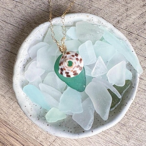 May include: A handcrafted necklace featuring a sea shell pendant with brown and white patterns, wire-wrapped on a piece of green sea glass. The pendant hangs from a delicate gold chain. The necklace rests on a bed of sea glass in a white ceramic bowl.